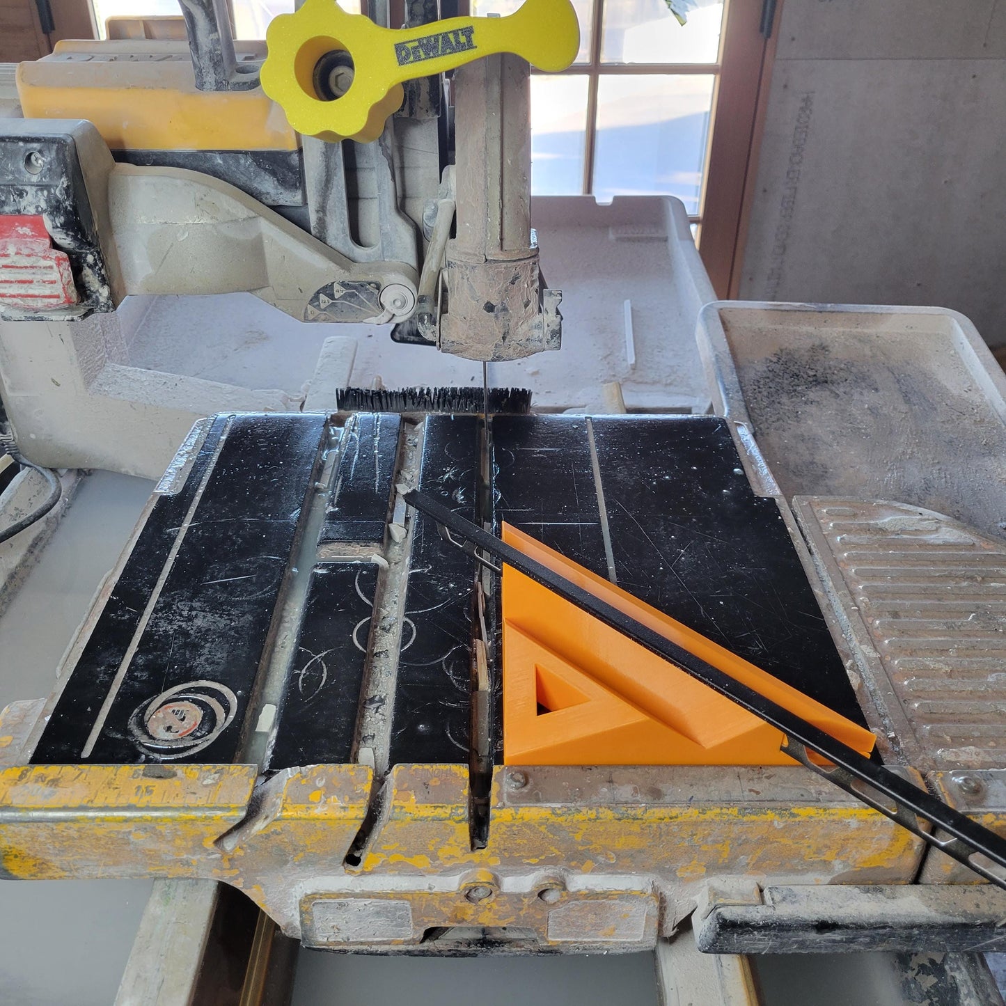 May include: A yellow and black tile saw with a black cutting surface and an orange angle guide. The saw has a yellow handle with the "DEWALT" logo. The saw is designed for cutting tiles.